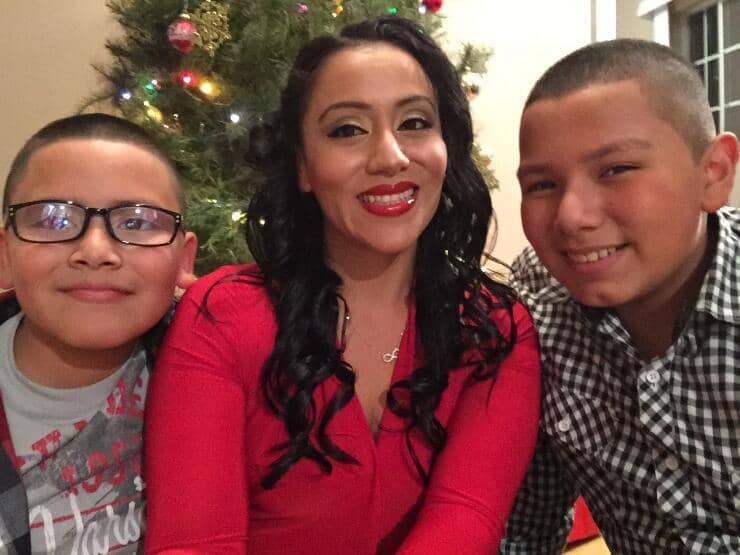 A family is in front of a christmas tree smiling at the camera. To the left is a young boy with buzzcut and glasses. In the middle is a woman with long black hair, red lipstick, natural makeup and a red shirt. To the right is a young teen with a toothy grin, he has a buzzcut and a black and white checkered shirt.